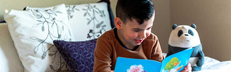 Boy reading a book with a stuffed animal panda in the background.